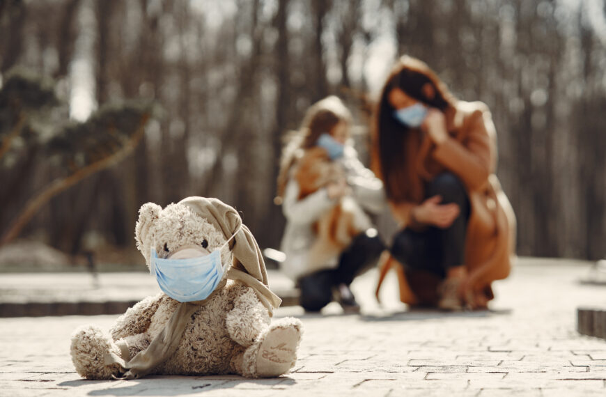 Shabby teddy bear in medical mask sitting on pavement against blurred mom and kid in park