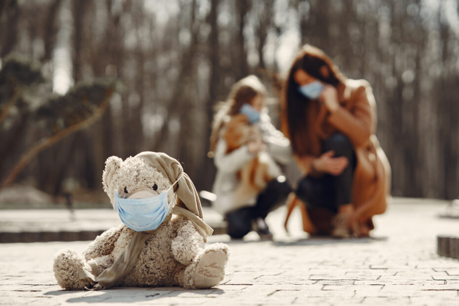 Shabby teddy bear in medical mask sitting on pavement against blurred mom and kid in park