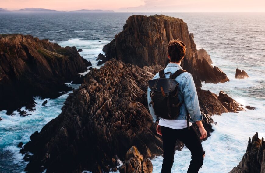 man standing near cliff looking at body of water during daytime